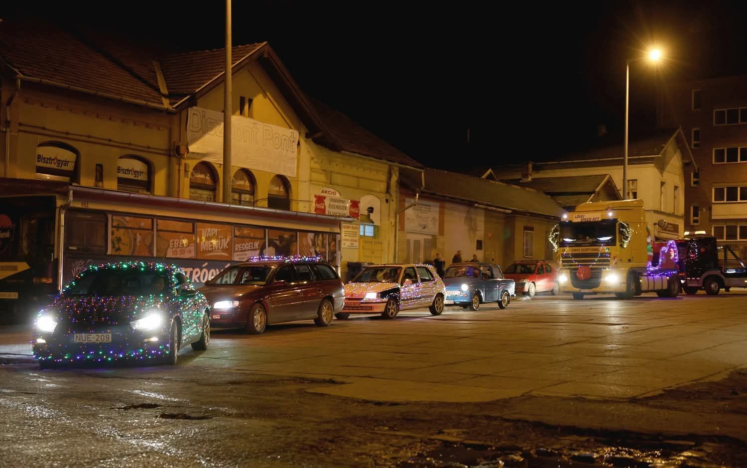 Fényfüzérekkel díszített autók konvoja vonul az esti belvárosban
